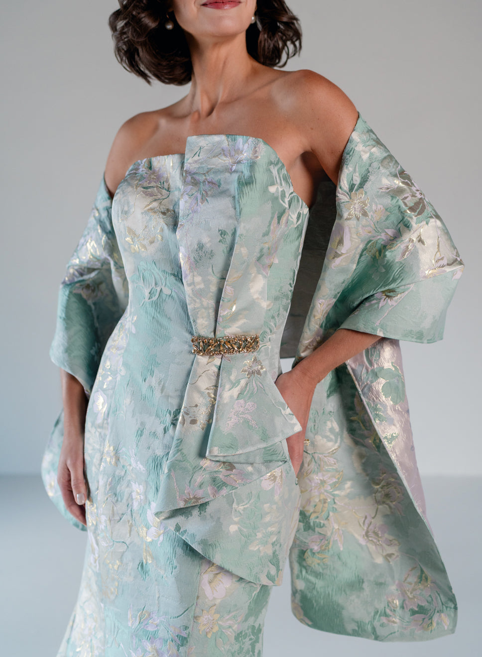 A woman models a strapless formal gown in a pale mint green and silver floral jacquard fabric, featuring dramatic draped cape sleeves and a jeweled waist detail, set against a neutral gray studio background. / Slim style in jacquard with straight neckline and large bow on side front decorated with small, beaded detail.