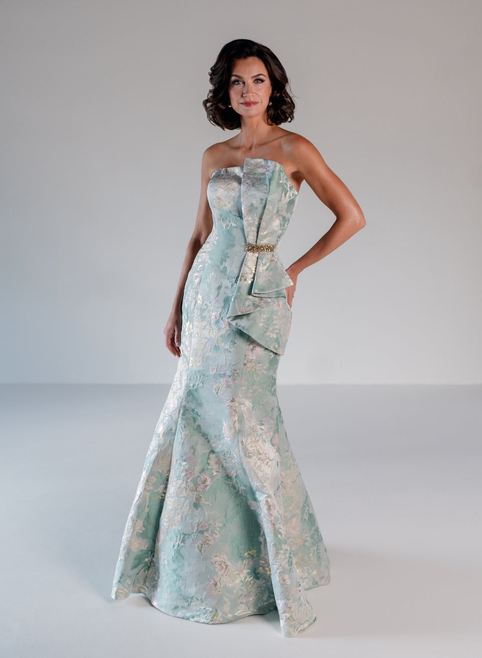 A woman models a strapless mermaid gown in a pale mint green and white floral brocade fabric, featuring a structured bodice with a draped bow detail and a gold waistband, set against a neutral studio background. / Slim style in jacquard with straight neckline and large bow on side front decorated with small, beaded detail.