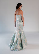 A woman stands in a studio wearing a strapless mermaid gown with a pale mint green and silver floral brocade pattern, photographed from the back against a light gray background. / Slim style in jacquard with straight neckline and large bow on side front decorated with small, beaded detail.
