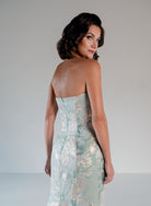 A woman wearing a strapless fitted gown with a pale mint green and white floral jacquard pattern, photographed from the back against a light gray studio background. / Slim style in jacquard with straight neckline and large bow on side front decorated with small, beaded detail.