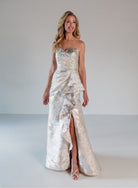 A woman modeling a strapless formal gown with a sweetheart neckline, ruched draped bodice, and a high-low ruffled hemline. The dress features a shimmering metallic silver and white jacquard pattern. / A-line brocade style with sweetheart neckline. Side draping on front dress finished with ruffle on side skirt area.