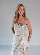 A woman modeling a strapless formal gown with a sweetheart neckline, featuring a textured champagne and silver metallic pattern with draped asymmetrical layers. / A-line brocade style with sweetheart neckline. Side draping on front dress finished with ruffle on side skirt area.