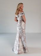 A woman modeling an off-the-shoulder floor-length gown with a metallic silver base and a rose-gold floral pattern, photographed in a studio setting. / A-line brocade style with sweetheart neckline. Side draping on front dress finished with ruffle on side skirt area.