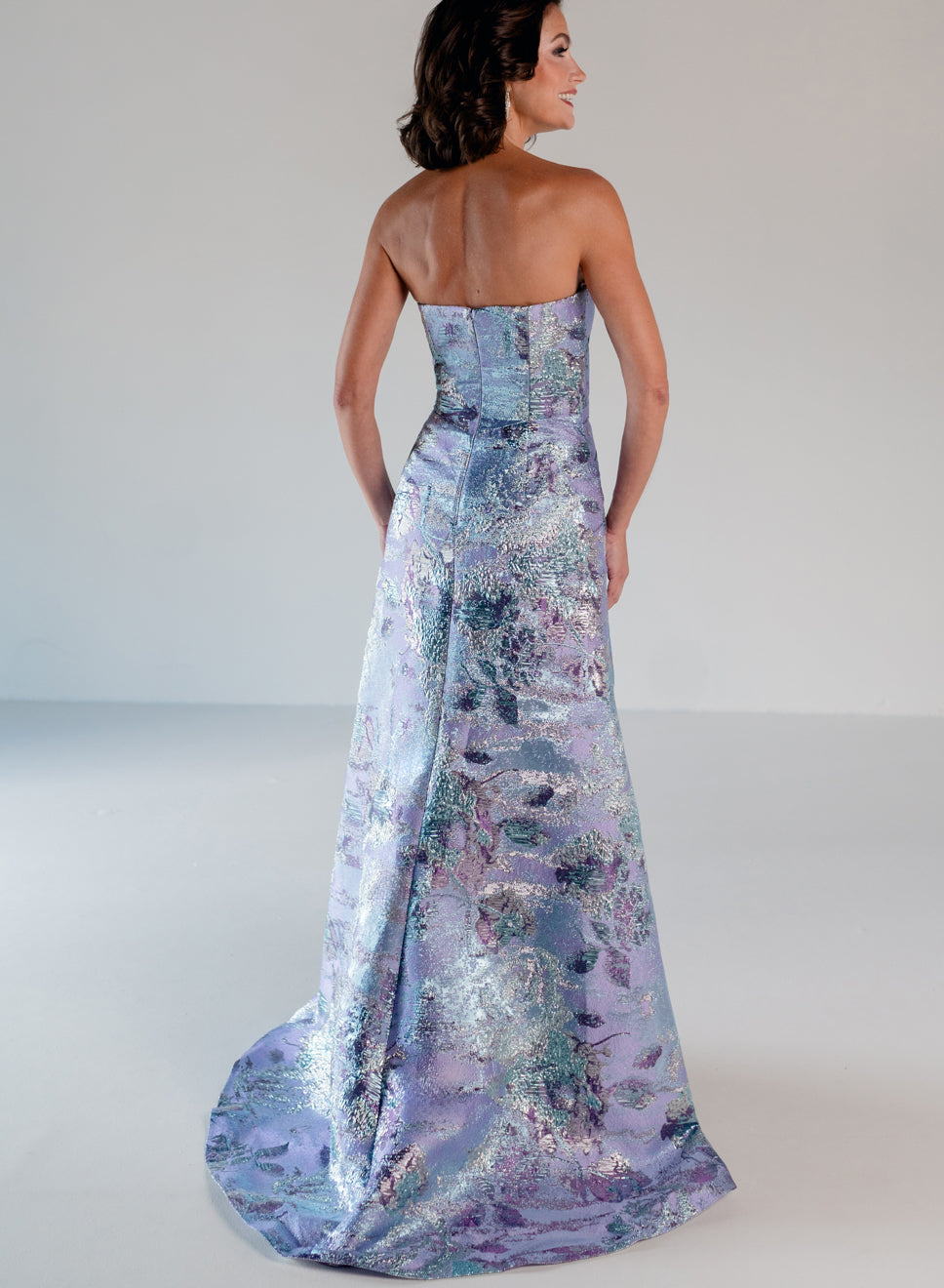 A woman modeling a strapless floor-length gown from the back, featuring a shimmering abstract pattern in lavender, blue, and silver tones against a plain light gray studio background. / A-line brocade style with sweetheart neckline. Side draping on front dress finished with ruffle on side skirt area.