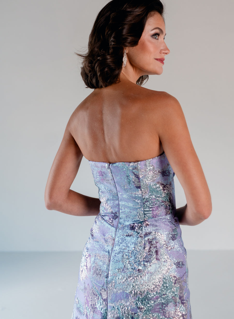 A woman shown from the back wearing a strapless evening gown with a shimmering lavender, blue, and silver patterned fabric against a light gray studio background. / A-line brocade style with sweetheart neckline. Side draping on front dress finished with ruffle on side skirt area.