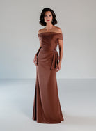 A woman modeling an off-the-shoulder floor-length gown in a warm brown shade, standing against a neutral light gray studio background. / A-line style in stretch satin with off-shoulder collar and side from draping finished with ruffle detail and beaded motif.