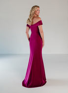A woman modeling a floor-length off-the-shoulder evening gown in rich magenta satin, photographed from the back against a clean light gray studio backdrop. / A-line style in stretch satin with off-shoulder collar and side from draping finished with ruffle detail and beaded motif.