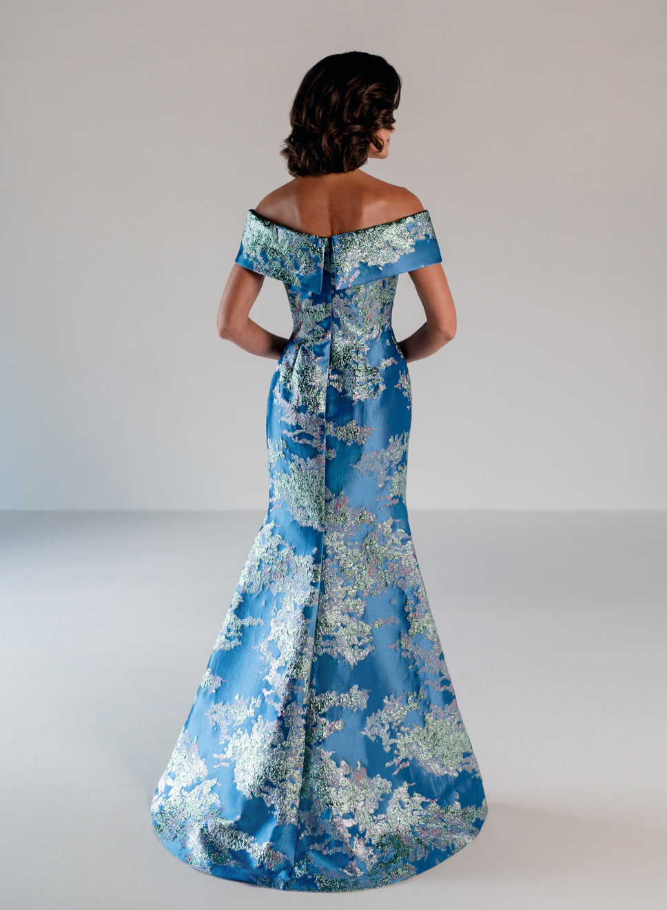 A woman viewed from the back wearing an off-the-shoulder blue satin gown with a white floral jacquard pattern, set against a plain light gray studio background. / Slim style in brocade with straight neckline and off-shoulder collar. Side draping on front dress.