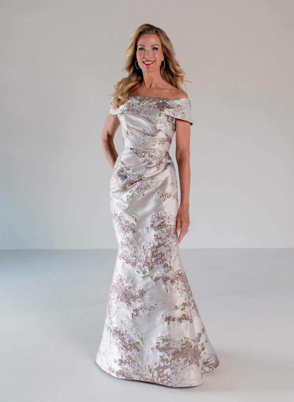 A woman modeling an off-the-shoulder floor-length gown with a white satin base and metallic rose gold floral pattern, set against a neutral light gray studio background. / Slim style in brocade with straight neckline and off-shoulder collar. Side draping on front dress.