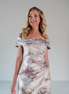 A woman modeling an off-the-shoulder champagne satin dress with a rose gold floral jacquard pattern, set against a neutral light gray studio background. / Slim style in brocade with straight neckline and off-shoulder collar. Side draping on front dress.