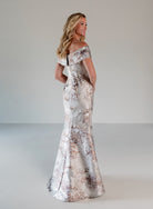 A woman modeling an off-the-shoulder floor-length gown with a shimmering silver base and a rose-gold floral pattern, photographed against a neutral light gray studio background. / Slim style in brocade with straight neckline and off-shoulder collar. Side draping on front dress.