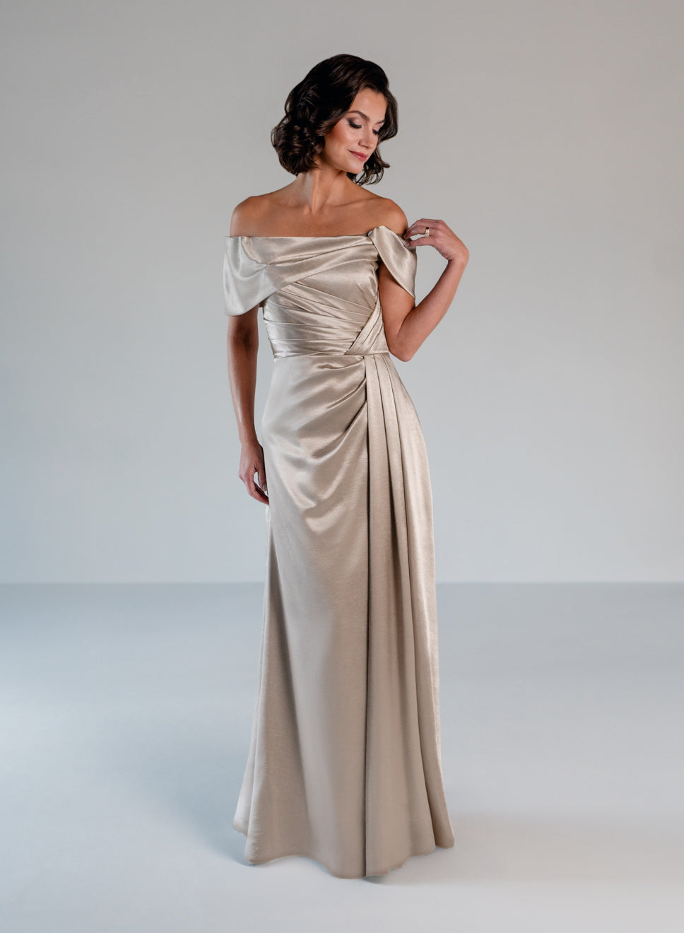 A woman modeling an off-the-shoulder floor-length satin gown in a champagne beige color, standing against a neutral light gray studio background. / Slim A-line style in smooth charmeuse with off-shoulder collar, asymmetrical pleating on front dress.