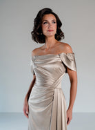 A woman modeling an off-the-shoulder champagne satin gown with draped detailing and a fitted waist, photographed against a neutral light gray studio background. / Slim A-line style in smooth charmeuse with off-shoulder collar, asymmetrical pleating on front dress.