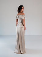 A woman modeling an off-the-shoulder floor-length satin gown in a champagne beige color, photographed against a neutral light gray studio background. / Slim A-line style in smooth charmeuse with off-shoulder collar, asymmetrical pleating on front dress.