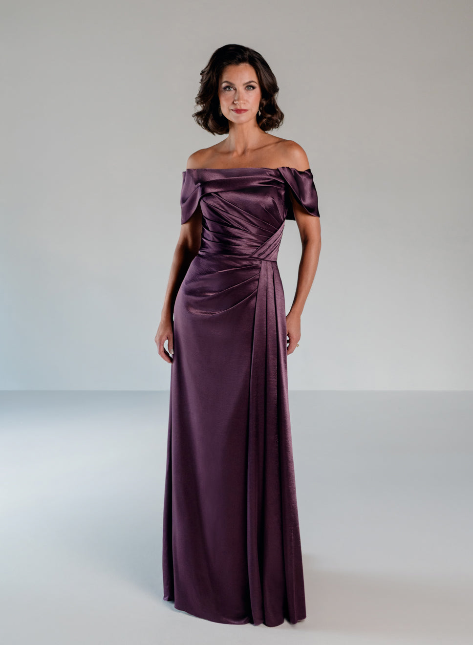 A woman modeling a floor-length off-the-shoulder gown in a rich plum purple satin fabric, set against a neutral light gray studio background. / Slim A-line style in smooth charmeuse with off-shoulder collar, asymmetrical pleating on front dress.