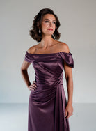 A woman modeling an off-the-shoulder, floor-length purple satin dress with draped detailing and a gathered waist, photographed against a light gray studio background. / Slim A-line style in smooth charmeuse with off-shoulder collar, asymmetrical pleating on front dress.