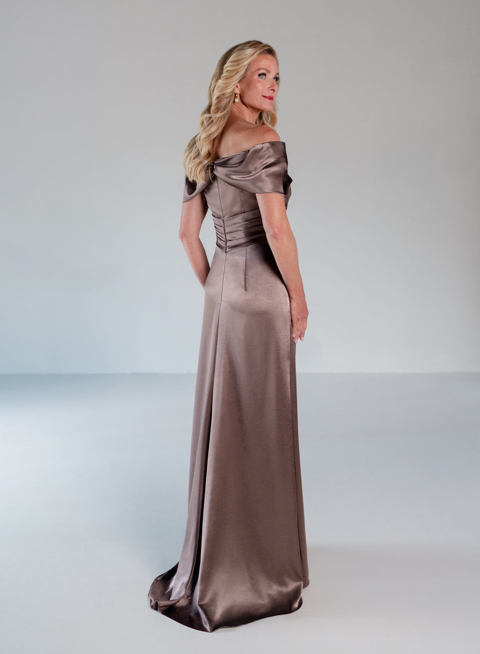 A woman modeling a floor-length off-the-shoulder satin gown in a taupe-brown color, photographed against a neutral light gray studio background. / Slim A-line style in smooth charmeuse with off-shoulder collar, asymmetrical pleating on front dress.