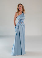 A woman modeling a floor-length light blue one-shoulder gown with a draped wrap-style skirt and blue floral embroidery near the waist, photographed in a clean studio setting. / Draped column gown with placed beading.