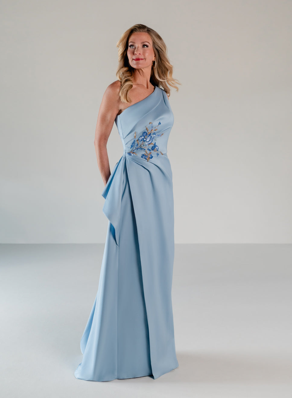 A woman modeling a floor-length light blue one-shoulder gown with a draped wrap-style skirt and blue floral embroidery near the waist, photographed in a clean studio setting. / Draped column gown with placed beading.