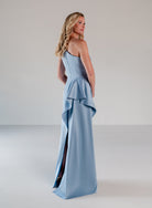 A woman modeling a light blue one-shoulder evening gown with a peplum waist and high side slit, photographed in a minimalist studio setting. / Draped column gown with placed beading.