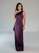 A woman modeling a floor-length, one-shoulder satin gown in deep plum purple. The dress features a structured bodice with a floral applique detail on the side and a draped, flowing skirt, set against a neutral light gray studio background. / Draped column gown with placed beading.