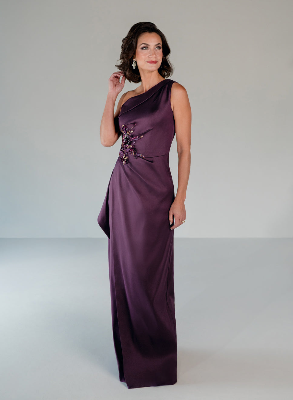 A woman modeling a floor-length, one-shoulder satin gown in deep plum purple. The dress features a structured bodice with a floral applique detail on the side and a draped, flowing skirt, set against a neutral light gray studio background. / Draped column gown with placed beading.