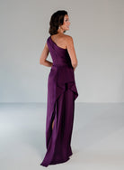 A woman modeling a floor-length one-shoulder gown in deep purple, shown from the back and side against a plain light gray studio background. / Draped column gown with placed beading.