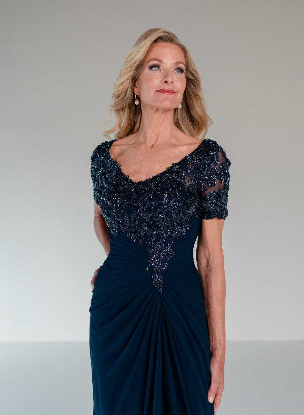 A woman modeling a floor-length navy evening gown with a heavily embellished dark beaded bodice, sheer short sleeves, and a draped gathered skirt against a plain light gray studio background. / Slim A-line style with chiffon skirt and beaded lace bodice, V-neck with short sleeves and side front draping on skirt.