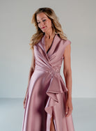A woman modeling a sleeveless dusty pink satin gown with a draped wrap-style neckline, ruffled skirt detail, and a jeweled embellished waist, photographed against a plain light gray studio background. / A-line mikado style with V-neck and short cup sleeves. Side front draping on bodice finished with ruffle on side front skirt.