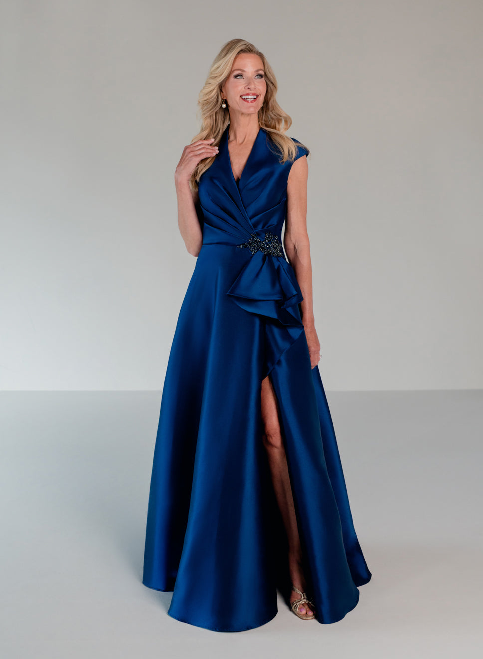 A woman modeling a floor-length royal blue satin gown with a wrap-style V-neck bodice, ruffled detailing at the waist, and a high front slit, photographed against a light gray studio background. / A-line mikado style with V-neck and short cup sleeves. Side front draping on bodice finished with ruffle on side front skirt.