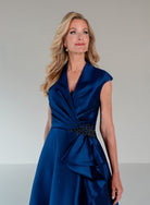 A woman modeling a sleeveless royal blue satin dress with a draped wrap-style neckline, gathered waist, and a dark embellished belt, photographed against a plain light gray studio background. / A-line mikado style with V-neck and short cup sleeves. Side front draping on bodice finished with ruffle on side front skirt.