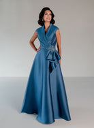 A woman modeling a floor-length blue satin gown with a wrap-style bodice, ruffled waist detail, and a jeweled embellishment at the waist, photographed against a plain light gray studio background. / A-line mikado style with V-neck and short cup sleeves. Side front draping on bodice finished with ruffle on side front skirt.