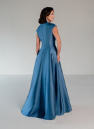 A woman modeling a floor-length satin gown from the back, showing a structured high neckline, cap sleeves, and a full skirt, in a dusty blue color against a neutral studio background. / A-line mikado style with V-neck and short cup sleeves. Side front draping on bodice finished with ruffle on side front skirt.