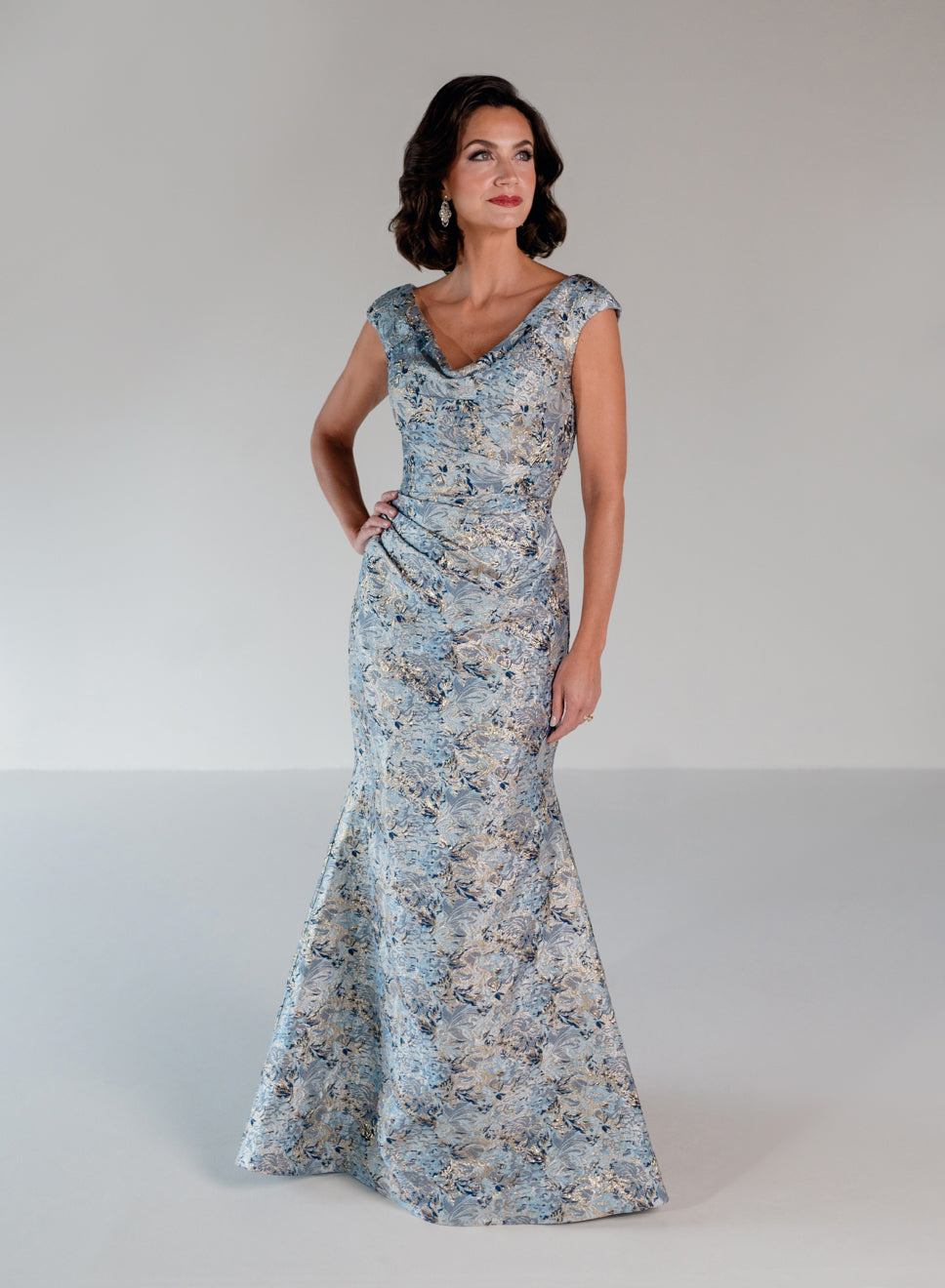 A woman modeling a floor-length, fitted evening gown with a blue and beige floral pattern against a neutral light gray studio background. / Slim brocade style with soft V-neck and small cup sleeves. Side draping on front dress.