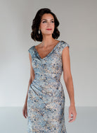 A woman modeling a sleeveless, V-neck dress with a blue, beige, and white abstract floral pattern, set against a plain light gray studio background. / Slim brocade style with soft V-neck and small cup sleeves. Side draping on front dress.