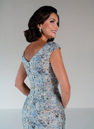 A woman wearing a fitted light blue sleeveless dress with a blue and beige abstract floral pattern, photographed against a plain light gray studio background. / Slim brocade style with soft V-neck and small cup sleeves. Side draping on front dress.