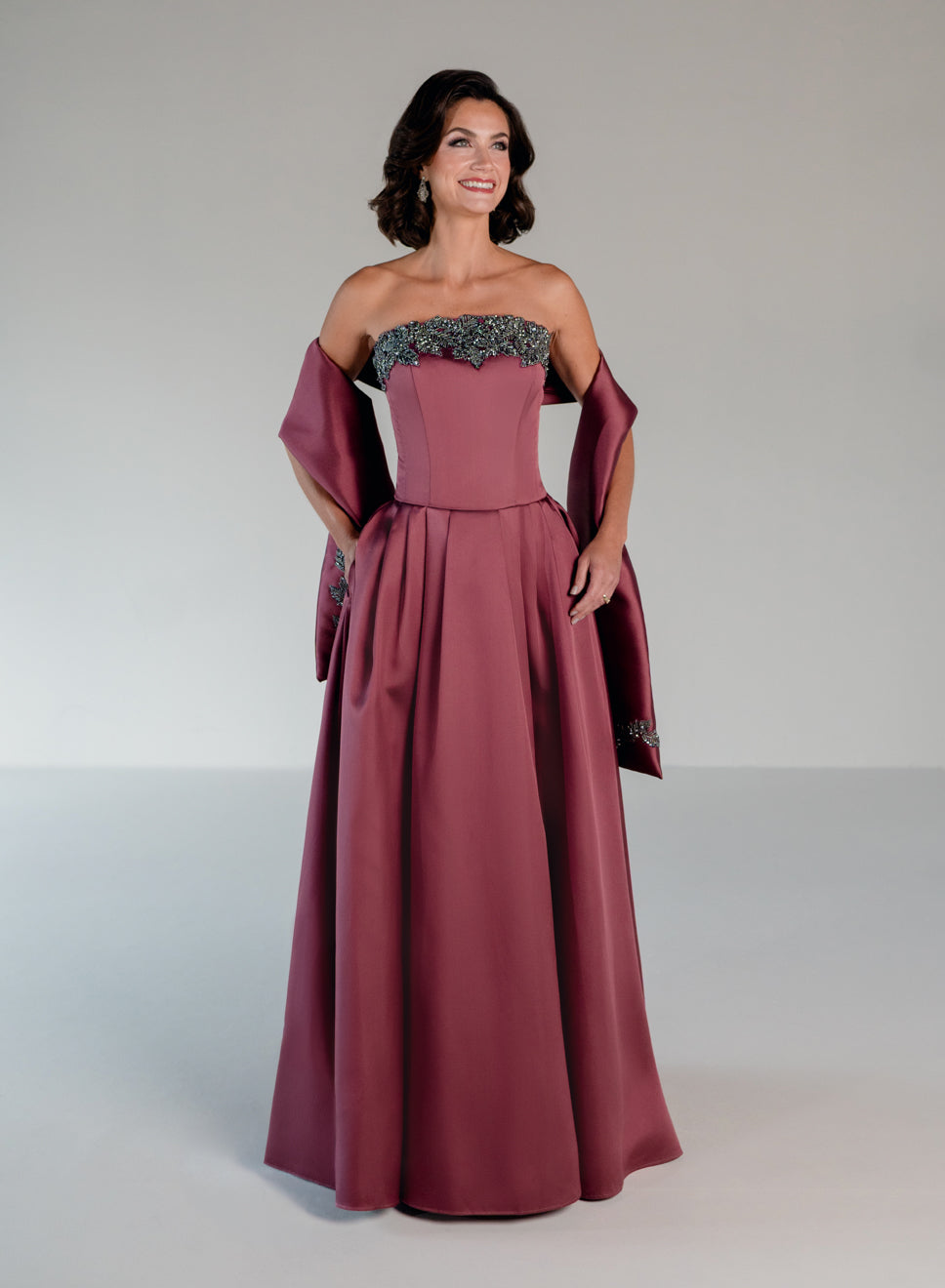 A woman modeling a floor-length off-the-shoulder gown in dusty rose with a dark embroidered lace bodice and matching draped sleeves, photographed against a neutral light gray studio background. / Mikado A-line ballgown with dropped waist and placed beading at the upward scoop neckline.