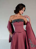 A woman wearing a strapless dusty rose evening gown with a structured corset bodice, a full skirt, and draped cape sleeves with silver beaded embellishments, posed against a neutral gray studio background. / Mikado A-line ballgown with dropped waist and placed beading at the upward scoop neckline.