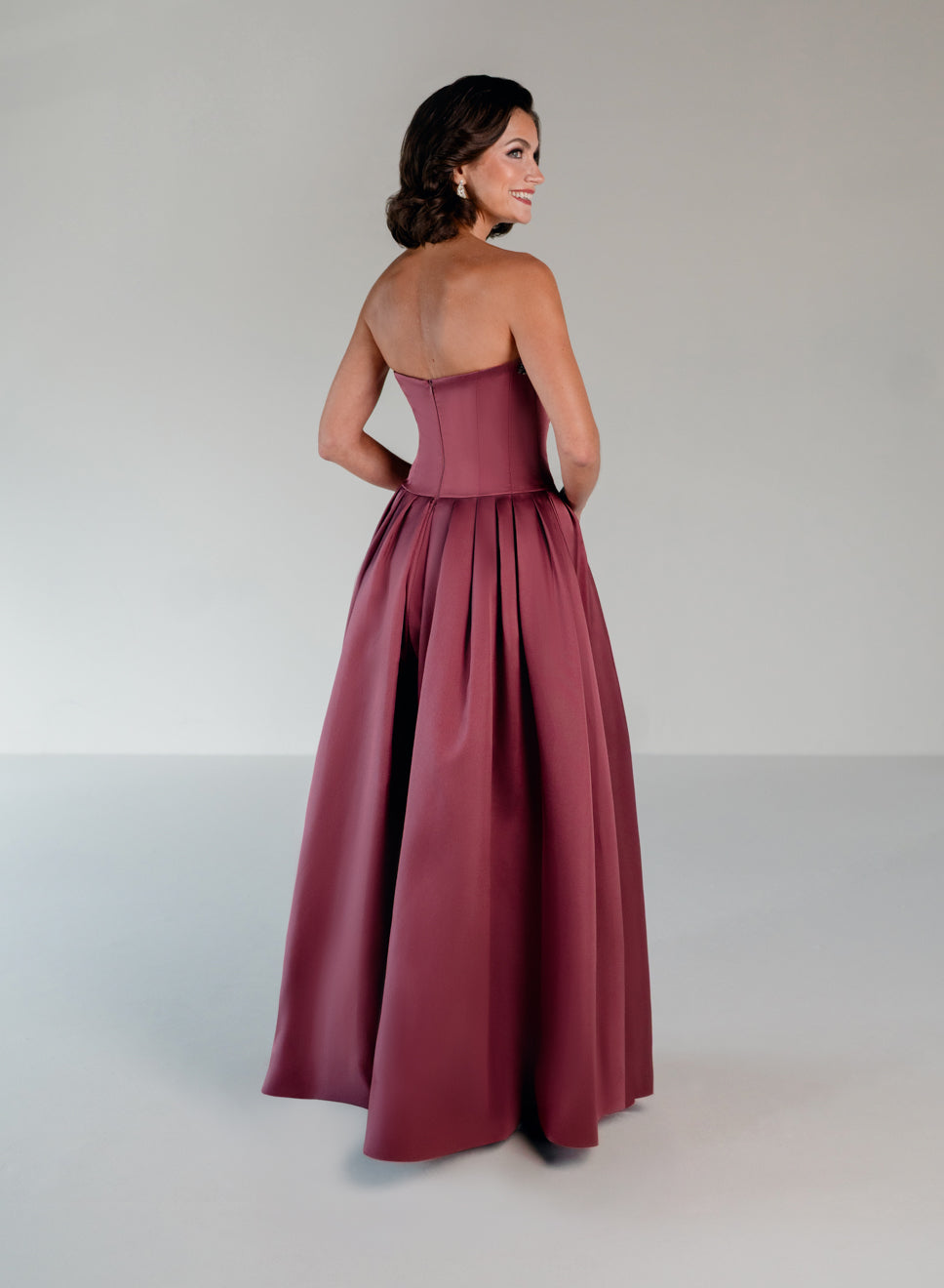 A woman modeling a strapless floor-length gown from the back, featuring a structured corset-style bodice and a full gathered skirt in a dusty rose or mauve shade, set against a plain light gray studio background. / Mikado A-line ballgown with dropped waist and placed beading at the upward scoop neckline.
