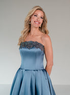 A woman modeling a strapless dusty blue satin gown with a heavily embellished beaded bodice against a neutral gray studio background.| Mikado A-line ballgown with dropped waist and placed beading at the upward scoop neckline.