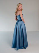 A woman modeling a strapless A-line formal gown in a dusty medium blue satin fabric, photographed against a neutral light gray studio background. / Mikado A-line ballgown with dropped waist and placed beading at the upward scoop neckline.