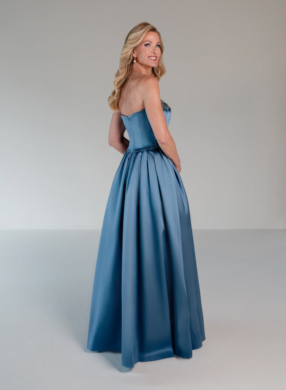 A woman modeling a strapless A-line formal gown in a dusty medium blue satin fabric, photographed against a neutral light gray studio background. / Mikado A-line ballgown with dropped waist and placed beading at the upward scoop neckline.