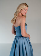 A blonde woman wearing a strapless dusty blue formal gown with a corseted bodice and full skirt, photographed from the back against a neutral light gray studio background. / Mikado A-line ballgown with dropped waist and placed beading at the upward scoop neckline.