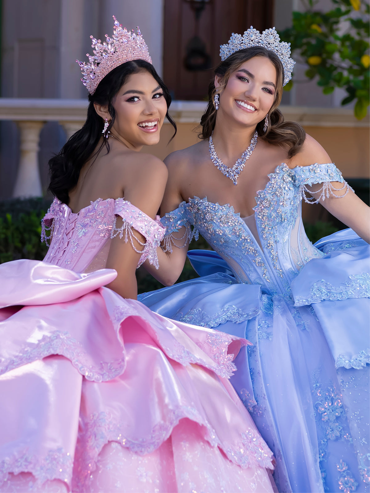 This gown features a fitted, embellished bodice with off-the-shoulder sleeves and a full tiered ball-gown skirt. Intricate appliqués decorate the layers, and a long flowing train with a large back bow completes the princess-style look.