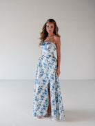 A woman modeling a strapless white maxi dress with a blue floral print and a high front slit, standing against a plain light gray studio background. / Straight neckline A-line gown with back bow detail.