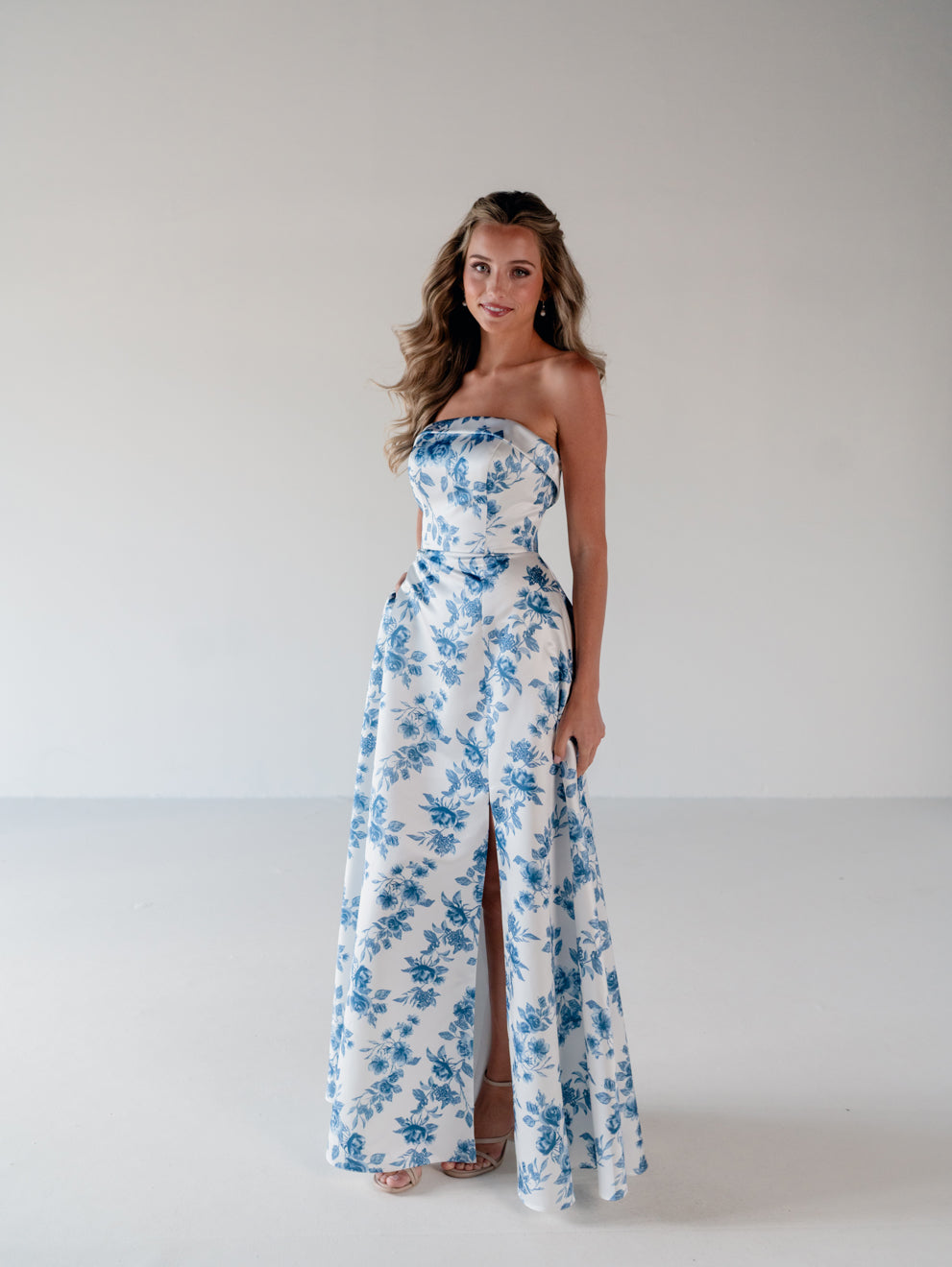 A woman modeling a strapless white maxi dress with a blue floral print and a high front slit, standing against a plain light gray studio background. / Straight neckline A-line gown with back bow detail.
