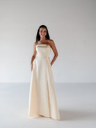 A woman modeling a strapless floor-length gown in a warm ivory or cream satin-like fabric, standing against a plain light gray studio background. / Straight neckline A-line gown with back bow detail.