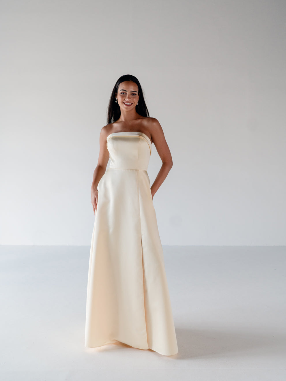 A woman modeling a strapless floor-length gown in a warm ivory or cream satin-like fabric, standing against a plain light gray studio background. / Straight neckline A-line gown with back bow detail.