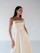 A woman modeling a strapless A-line gown in a warm ivory or cream satin-like fabric against a plain light gray studio background. / Straight neckline A-line gown with back bow detail.