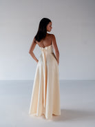 A woman stands with her back to the camera wearing a strapless cream satin gown with a large bow at the back of the neckline and a flowing floor-length skirt, photographed in a minimalist studio setting. / Straight neckline A-line gown with back bow detail.